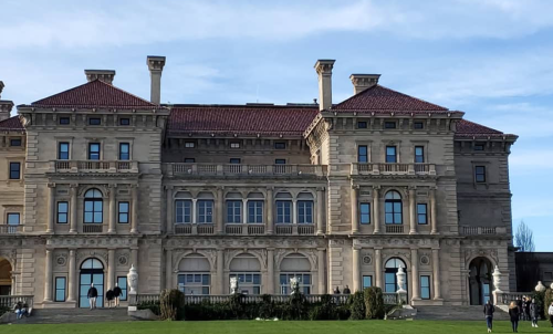 A grand mansion with a red-tiled roof, ornate architecture, and a manicured lawn under a clear blue sky.