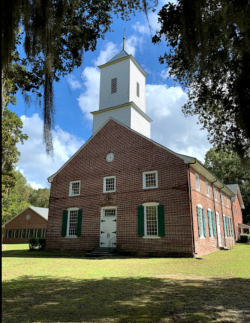 Historic brick church with a tall white steeple, green shutters, and a grassy area surrounded by trees.