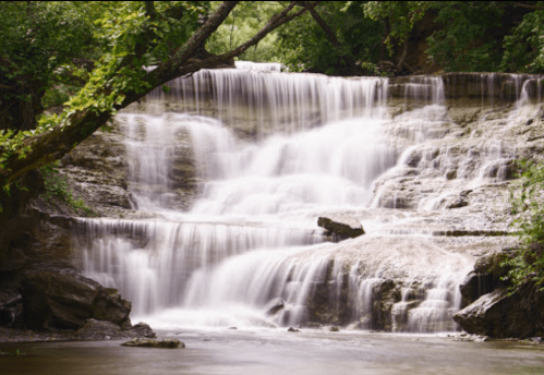 A serene waterfall cascading over rocky steps, surrounded by lush green trees and a calm river below.