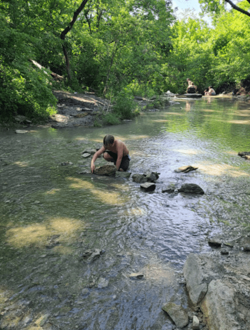 A person crouches by a shallow stream, surrounded by greenery, while others relax on rocks in the background.