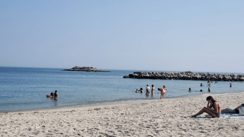 A sunny beach scene with people swimming and relaxing on the sand, and rocky formations in the background.