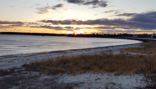 A serene beach at sunset, with gentle waves and a sandy shore, framed by clouds and distant buildings.