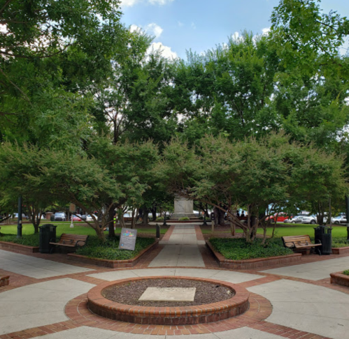 A park with green trees, benches, and a circular pathway leading to a central area with a monument.