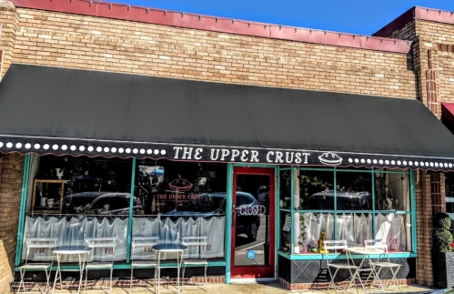 A brick storefront with a black awning reading "The Upper Crust," featuring outdoor tables and a closed sign.