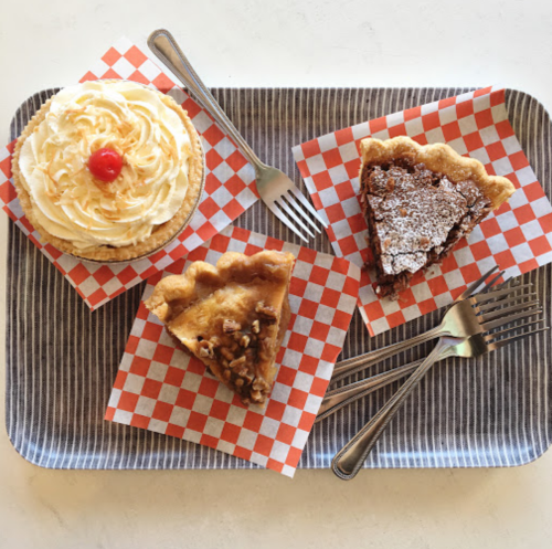 A tray with three dessert slices: a cream pie with a cherry, a chocolate pie, and a pecan pie, all on checkered paper.