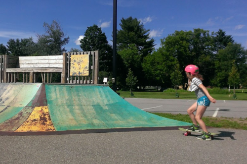 A girl in a pink helmet skateboards down a ramp at a skate park on a sunny day.