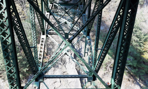 View from beneath a green metal bridge, showcasing intricate steel beams and rocky terrain below.