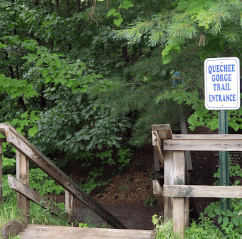 Sign for Quechee Gorge Trail entrance with wooden steps surrounded by lush greenery.