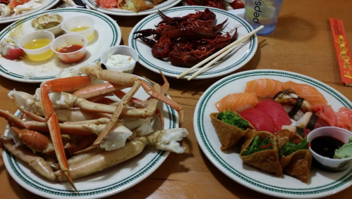 A table spread with plates of sushi, crab legs, crawfish, and dipping sauces.