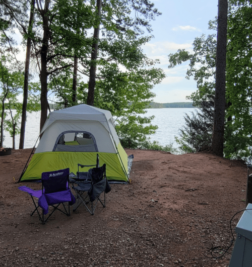 A green and white tent set up by a lake, surrounded by trees and two camping chairs on a gravel area.