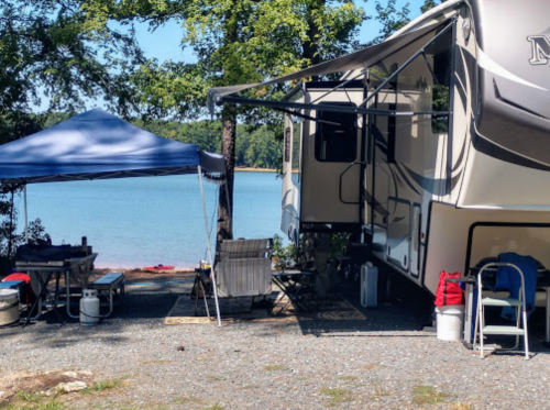 A campsite by a lake featuring an RV, a blue canopy, chairs, and a grill under a sunny sky.