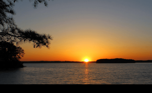 Sunset over a calm lake, with silhouettes of trees and islands against a vibrant orange and blue sky.
