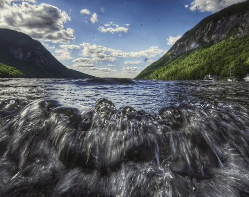 Waves gently lap at the shore, framed by lush green mountains and a blue sky with fluffy clouds.