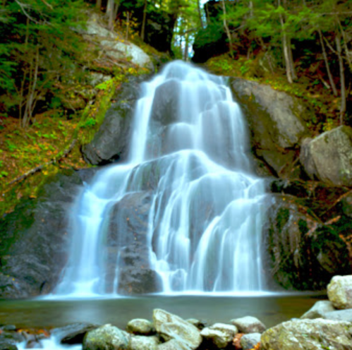 A serene waterfall cascading over rocks, surrounded by lush green trees and a tranquil pool at the base.