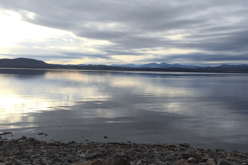 A calm lake reflects the cloudy sky, surrounded by distant mountains and rocky shoreline.