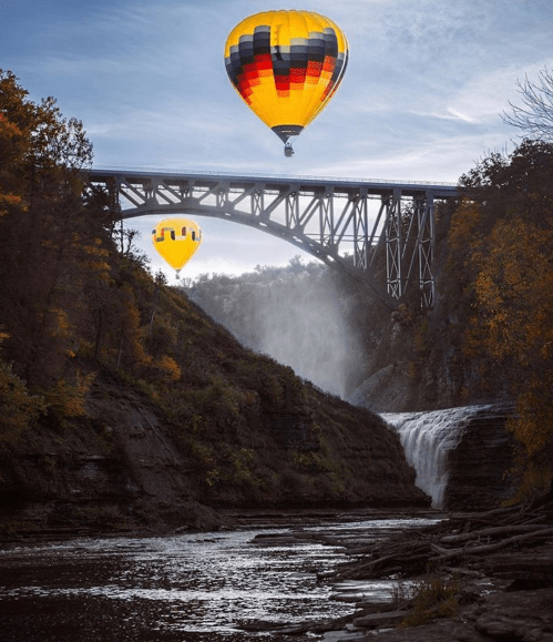 Two colorful hot air balloons float above a waterfall and bridge, surrounded by autumn foliage.