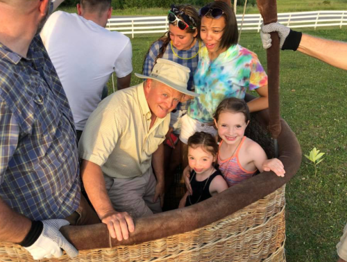 A group of people, including children, gather around a hot air balloon basket, smiling and enjoying the moment outdoors.