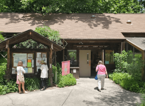 Visitors approach a rustic building surrounded by greenery, with informational signs and a colorful banner.