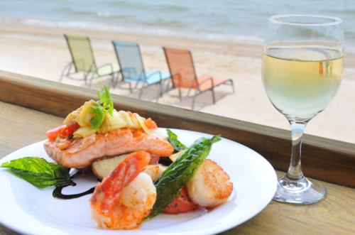 A plate of salmon, shrimp, and scallops with a glass of white wine, overlooking a beach with colorful chairs.