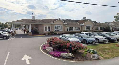 A brick building with large windows, surrounded by flowers and parked cars, under a cloudy sky.