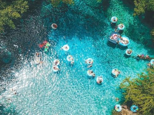 Aerial view of a clear blue water area with people floating on inner tubes and relaxing among trees.