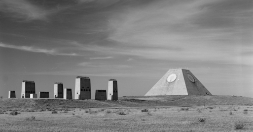 A large pyramid-shaped structure with circular features, surrounded by several rectangular concrete blocks on a grassy landscape.