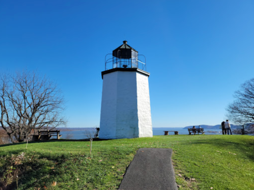 A white lighthouse stands on a grassy hill, overlooking a river under a clear blue sky, with benches nearby.