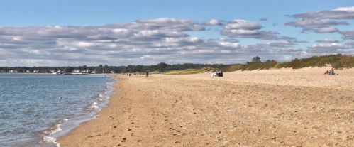 A sandy beach with gentle waves, dotted with people enjoying the sunny day under a partly cloudy sky.