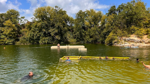 People swimming and relaxing in a green lake surrounded by trees, with a floating platform in the background.