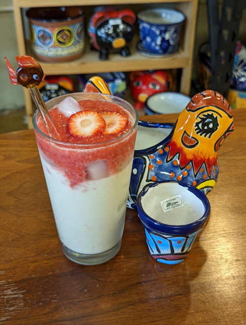 A tall glass of layered strawberry and cream drink, garnished with strawberries, next to colorful ceramic decorations.