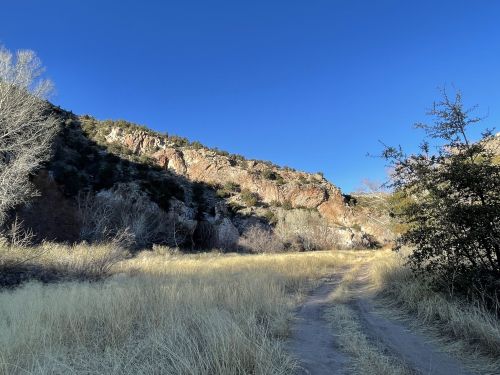 A dirt path leads through tall grass, flanked by rocky cliffs and clear blue skies.