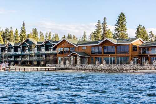 Lakeside view of a rustic lodge with large windows, surrounded by trees and a calm water surface.