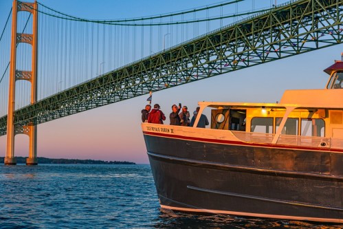 A boat with passengers sails under a large green bridge at sunset, reflecting warm colors on the water.