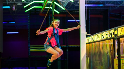 A girl in a bright shirt swings on a zip line in a colorful indoor adventure park.