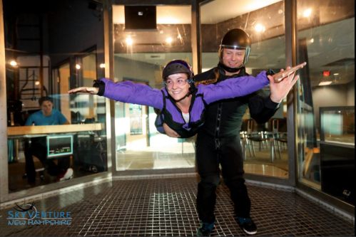 A person in a purple jumpsuit is flying in a wind tunnel, supported by an instructor in a black suit.