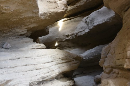 Sunlight filters through rocky crevices in a narrow canyon, highlighting textured, sandy walls.