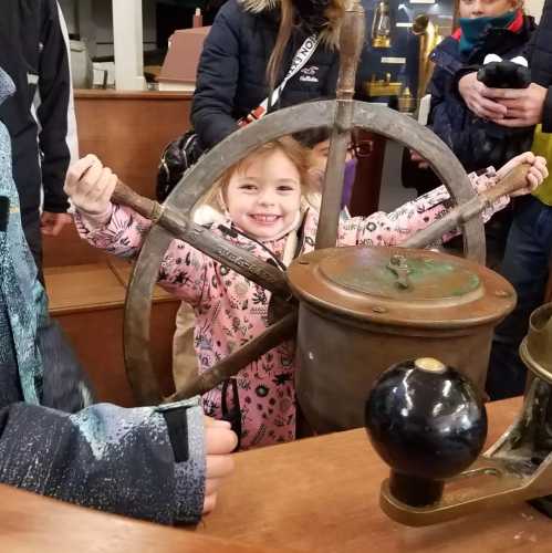 A young girl smiles while holding a ship's steering wheel, surrounded by other children in a museum setting.