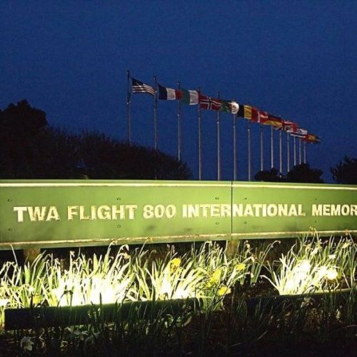 TWA Flight 800 International Memorial sign illuminated at night, surrounded by flags and greenery.