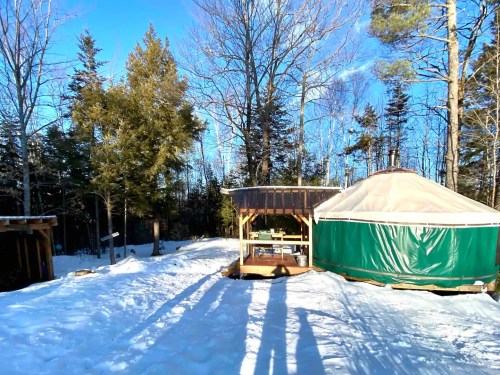 A snowy landscape featuring a yurt and a wooden structure, surrounded by trees under a clear blue sky.