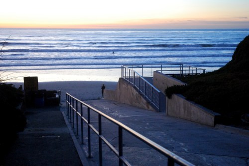 A pathway leads down to a beach at sunset, with gentle waves and a colorful sky in the background.