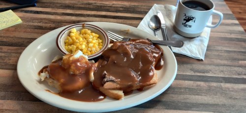 A plate of food with mashed potatoes, gravy, corn, and sliced meat, alongside a cup of coffee on a wooden table.