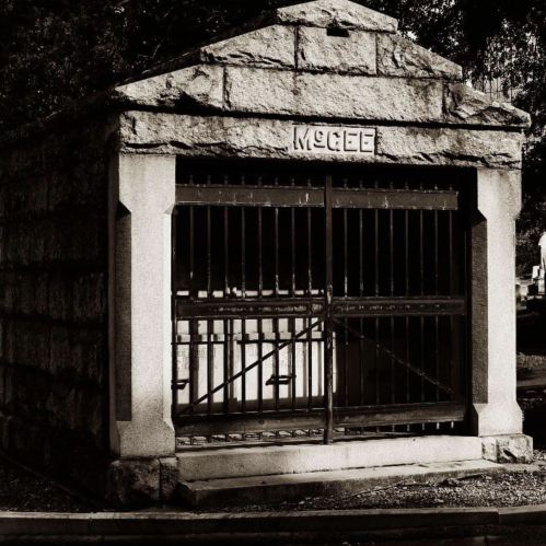 A stone mausoleum with barred gates, labeled "McGee," set in a cemetery. The image is in black and white.