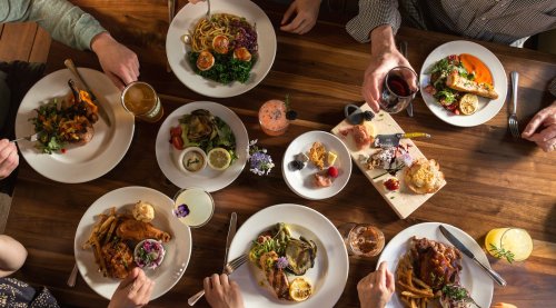 A table filled with various dishes, drinks, and hands reaching for food, creating a vibrant dining experience.