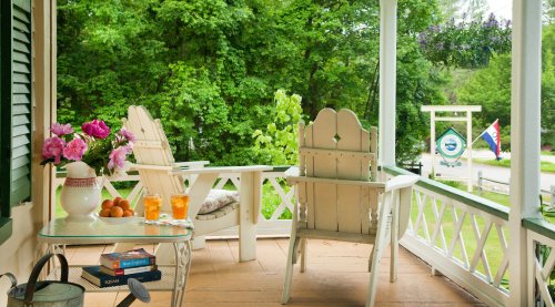 A cozy porch with white chairs, a small table with drinks and books, surrounded by lush greenery.