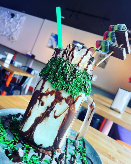 A colorful milkshake topped with whipped cream, chocolate drizzle, and green sprinkles, served on a plate with chocolate chips.