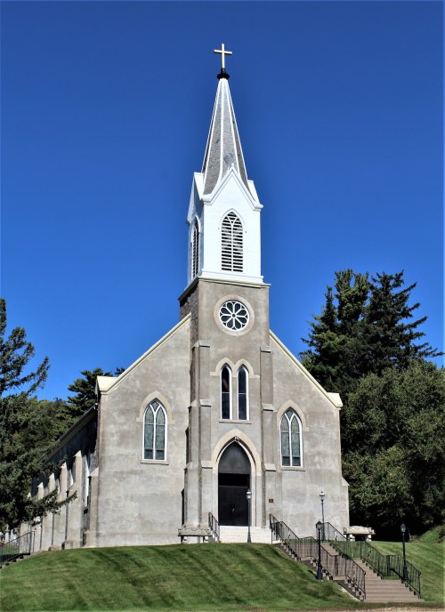 A tall church with a steeple and cross, surrounded by trees and a clear blue sky. Steps lead to the entrance.
