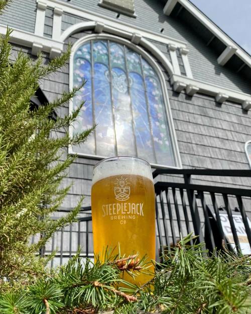 A glass of beer sits in front of a building with a stained glass window, surrounded by greenery.