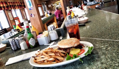 A burger with fries on a plate at a diner, with a drink and condiments on the counter in the background.