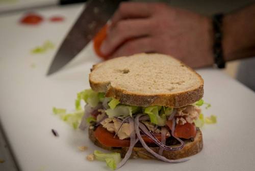A close-up of a sandwich with tuna, lettuce, tomato, and onion, with a hand slicing a tomato in the background.