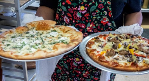 A person in an apron holds two pizzas: one with white sauce and greens, the other with meat and colorful toppings.